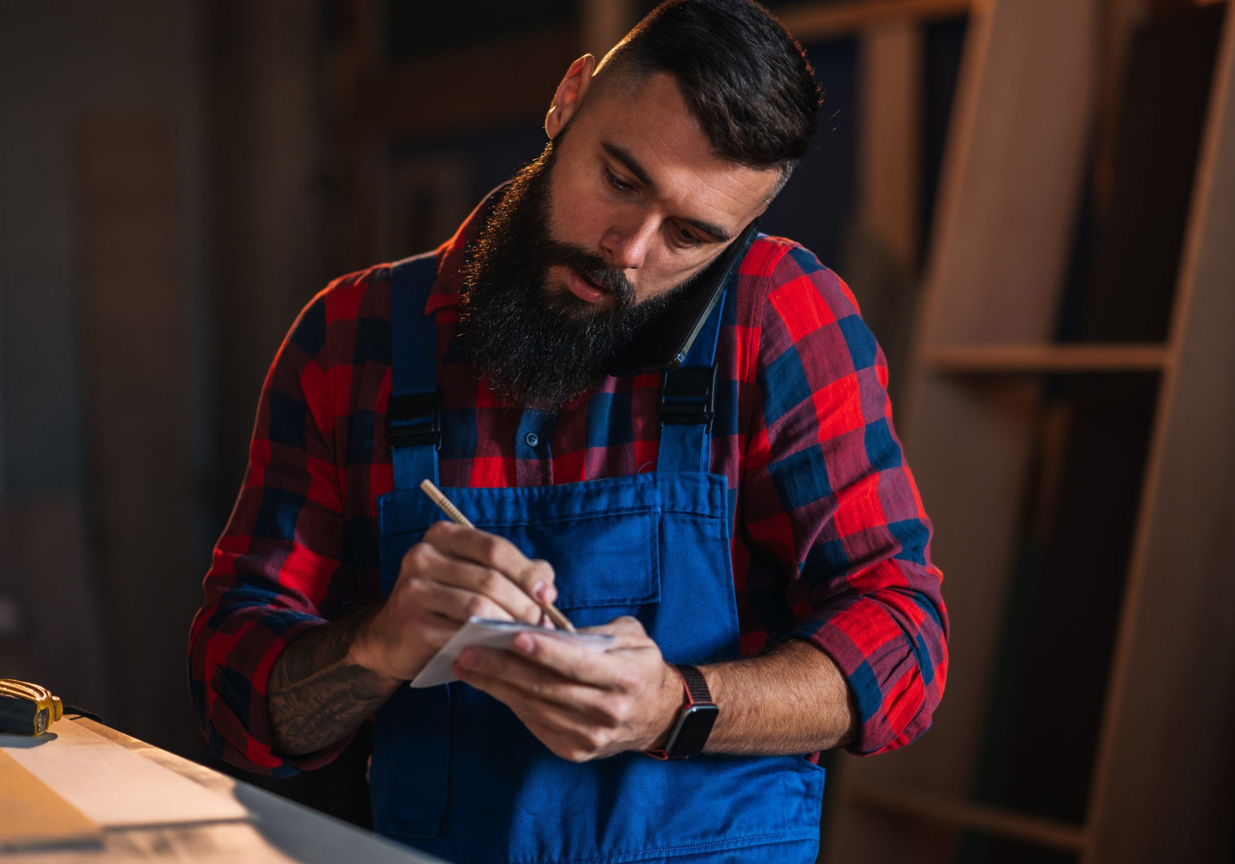 Young male carpenter working in workshop and using smartphone Young male carpenter working in workshop and using smartphone while talking with a client
